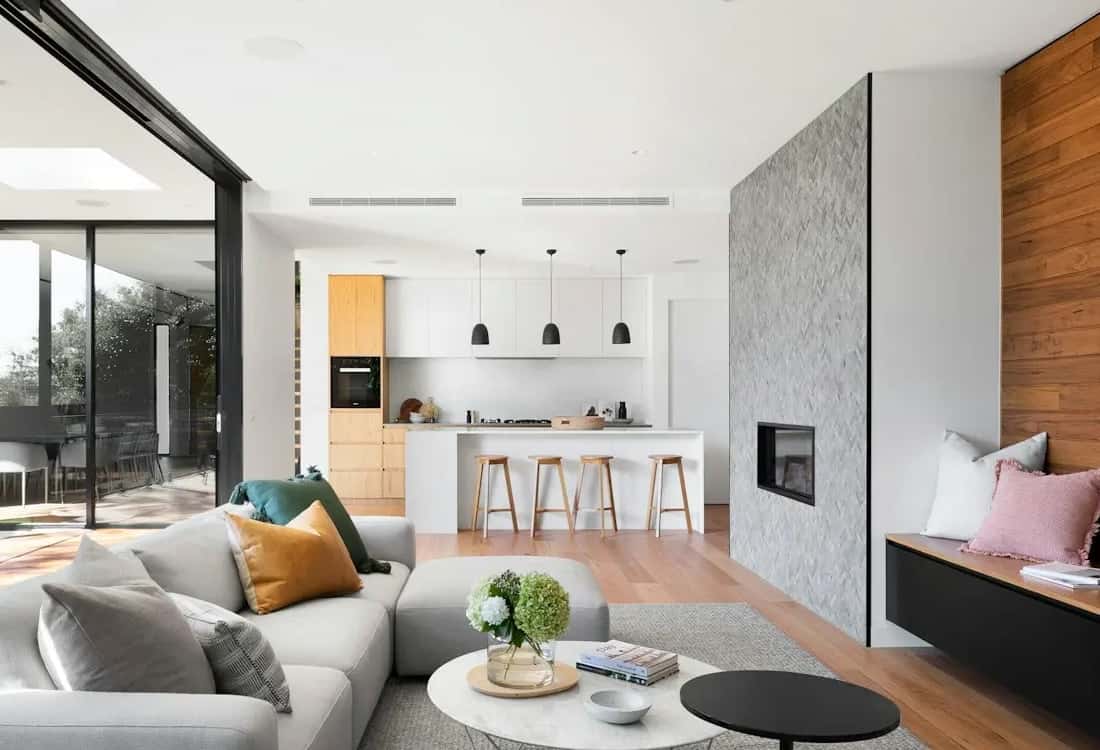 Open-plan kitchen with a white island, timber bar stools and black pendant lights, seen from the living area