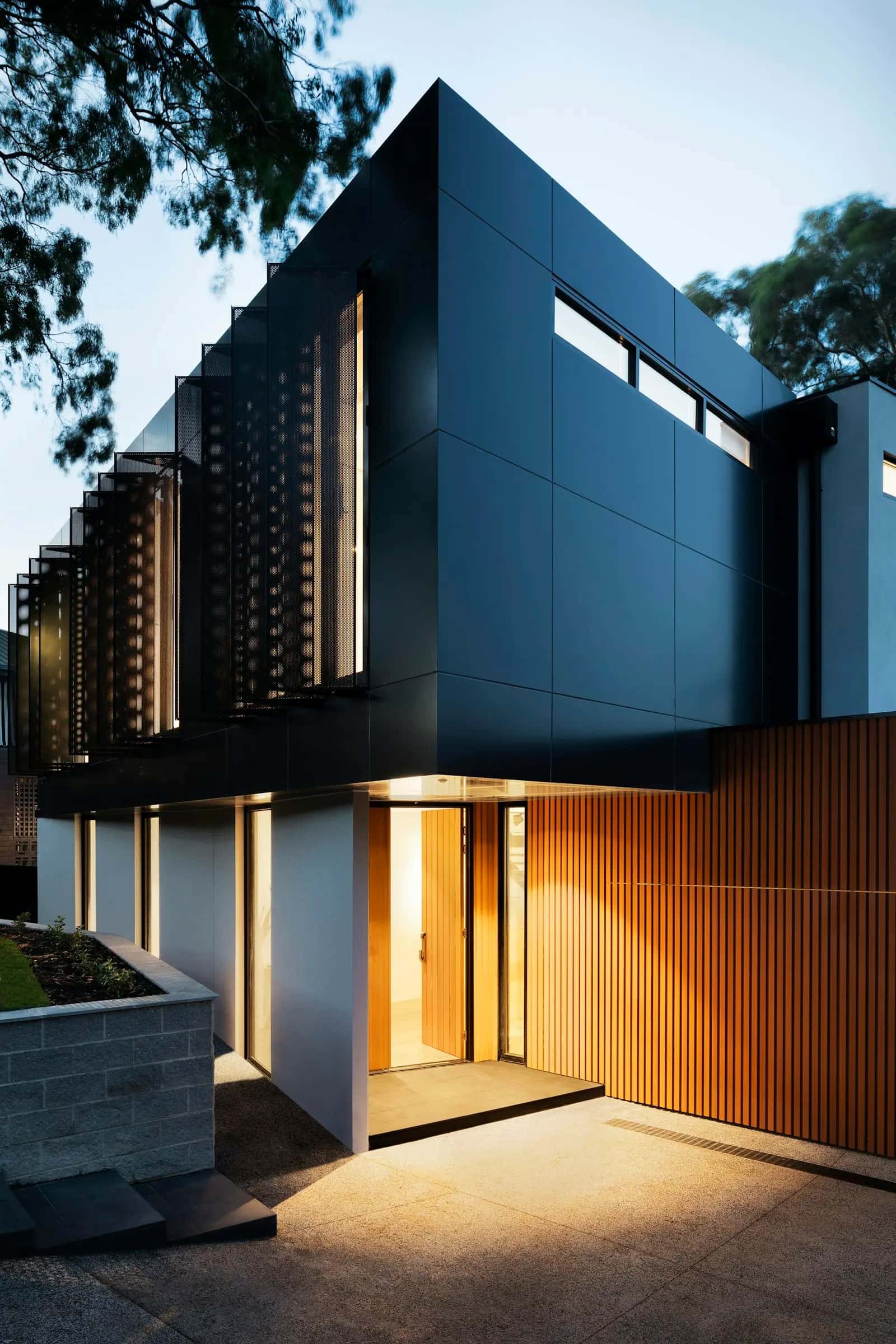 Modern two-story home at dusk with dark cladding, a timber-slat wall and a lit front entry